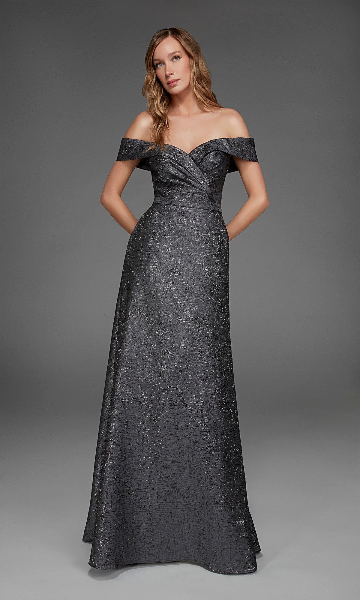 A woman stands in a studio wearing the Long Formal Dress: Alyce 27882—a floor-length, dark metallic jacquard evening gown featuring textured fabric and discreet pockets.